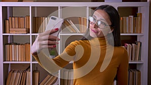 Beautiful brunette female teacher in glasses making funny selfies on smartphone showing tongue joyfully at the library.