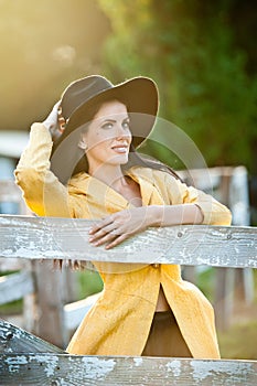 Beautiful brunette girl with country look near an old wooden fence. Attractive woman with black hat and yellow coat