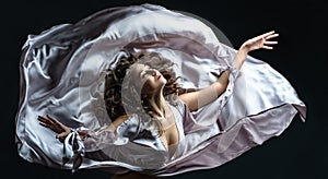Beautiful brunette girl with curly hair in the darkness and light in silver satin flying dress awesome poses in dance