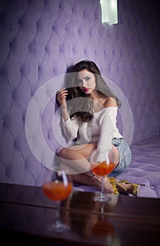 Beautiful brunette girl in denim shorts and white blouse posing on lilac textured background with two drink glasses foreground