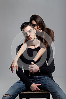 Beautiful brunette girl hugging from behind a guy dressed in a black long sleeve t-shirt and jeans sitting on a wooden