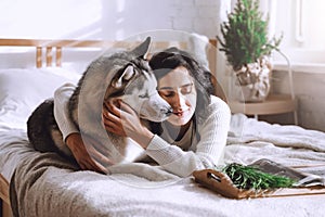 A beautiful brunette girl lies on the bed with a white husky dog at home. Sunny morning