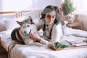 A beautiful brunette girl lies on the bed with a white husky dog at home. Sunny morning