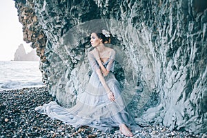 Beautiful brunette girl in a long grey dress with a veil sitting by the sea