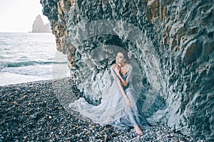 Beautiful brunette girl in a long grey dress with a veil sitting by the sea, near the cliff,