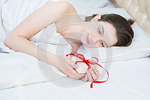 Beautiful brunette girl lying on the bed holding a white gift box with a red ribbon at home, front view, copy space