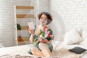 a beautiful brunette girl sits on a bed and holds a card and a bouquet of tulips in her hands. snow-white smile