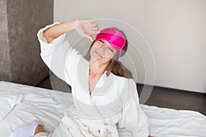 Beautiful brunette girl stretching in bed in a mask for sleeping. She just woke up. Morning Concept.