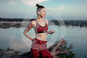 A beautiful brunette girl in stylish sports clothes is resting after a sport active workout near the lake. Listens to music on hea