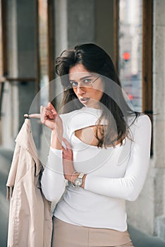 beautiful brunette girl in a sunglasses dressed in white blouse with a beige coat in hands. Stylish trendy fashion