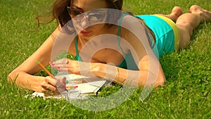 Beautiful brunette girl in sunglasses laying on the grass and colorizing. 4K steadicam shot, vivid colors