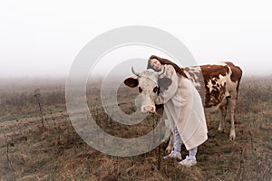 Beautiful brunette girl in white coat stands among the meadow in fog with red and white coe, hugging her and looking at