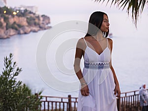 Beautiful brunette girl in a white dress waiting near sea at sunset