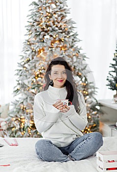 A beautiful brunette girl in a white sweater and blue jeans sits on a bed with a cup of hot drink in her hands