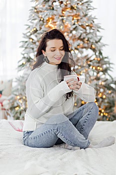 A beautiful brunette girl in a white sweater and blue jeans sits on a bed with a cup of hot drink in her hands