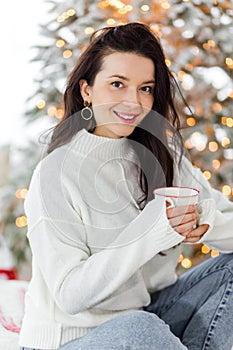 A beautiful brunette girl in a white sweater and blue jeans sits on a bed with a cup of hot drink in her hands