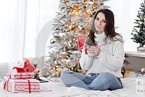 A beautiful brunette girl in a white sweater and blue jeans sits on a bed with a cup of hot drink in her hands