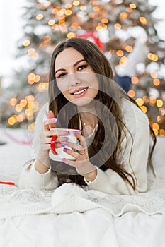 A beautiful brunette girl in a white sweater and blue jeans sits on a bed with a cup of hot drink in her hands.