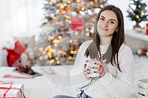 A beautiful brunette girl in a white sweater and blue jeans sits on a bed with a cup of hot drink in her hands