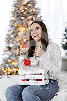 A beautiful brunette girl in a white sweater sits on a bed against the background of Christmas lights and holds red