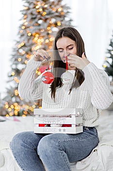 A beautiful brunette girl in a white sweater sits on a bed against the background of Christmas lights and holds red