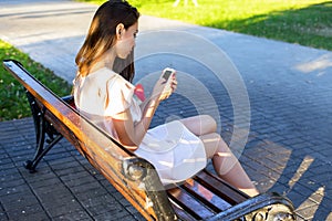 Beautiful brunette girl writing a message phone park sitting on bench in dress, summer day business woman relaxing