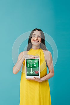 Brunette girl in yellow dress showing digital tablet with cardiological app isolated on turquoise photo