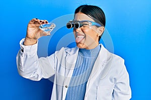 Beautiful brunette jeweller woman holding brilliant diamond stone wearing magnifier glasses sticking tongue out happy with funny