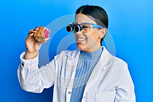 Beautiful brunette jeweller woman holding geode stone wearing magnifier glasses winking looking at the camera with sexy