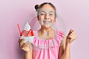 Beautiful brunette little girl eating strawberry ice cream screaming proud, celebrating victory and success very excited with
