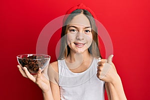 Beautiful brunette little girl holding bowl of raisins smiling happy and positive, thumb up doing excellent and approval sign