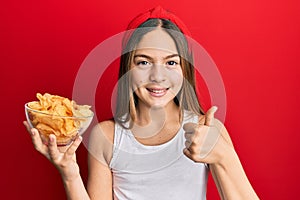 Beautiful brunette little girl holding potato chips smiling happy and positive, thumb up doing excellent and approval sign