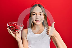 Beautiful brunette little girl holding red pepper smiling happy and positive, thumb up doing excellent and approval sign