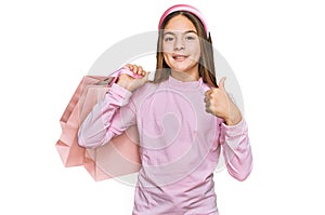 Beautiful brunette little girl holding shopping bags smiling happy and positive, thumb up doing excellent and approval sign