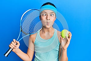 Beautiful brunette little girl playing tennis holding racket and ball making fish face with mouth and squinting eyes, crazy and