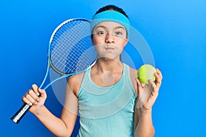 Beautiful brunette little girl playing tennis holding racket and ball puffing cheeks with funny face