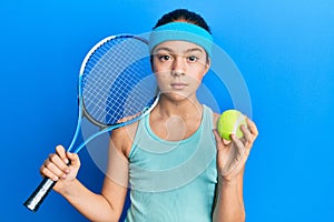 Beautiful brunette little girl playing tennis holding racket and ball relaxed with serious expression on face