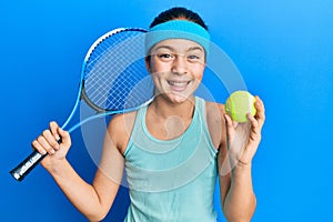 Beautiful brunette little girl playing tennis holding racket and ball smiling and laughing hard out loud because funny crazy joke