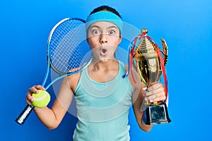 Beautiful brunette little girl playing tennis holding trophy afraid and shocked with surprise and amazed expression, fear and