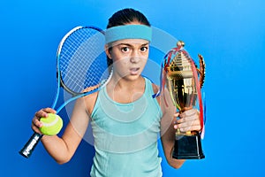 Beautiful brunette little girl playing tennis holding trophy clueless and confused expression