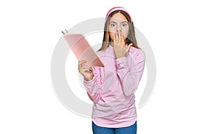 Beautiful brunette little girl reading a book covering mouth with hand, shocked and afraid for mistake