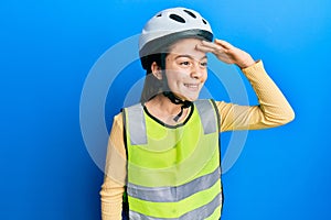 Beautiful brunette little girl wearing bike helmet and reflective vest very happy and smiling looking far away with hand over head