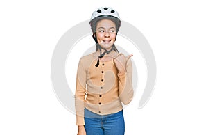 Beautiful brunette little girl wearing bike helmet smiling with happy face looking and pointing to the side with thumb up