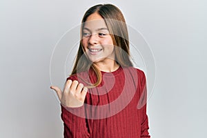 Beautiful brunette little girl wearing casual sweater pointing thumb up to the side smiling happy with open mouth