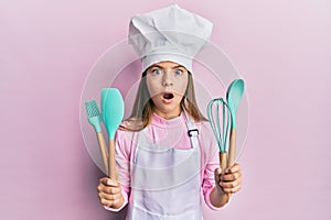 Beautiful brunette little girl wearing professional cook apron holding cooking tools afraid and shocked with surprise and amazed