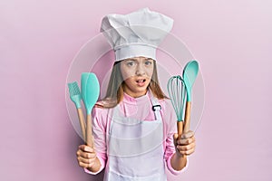 Beautiful brunette little girl wearing professional cook apron holding cooking tools clueless and confused expression