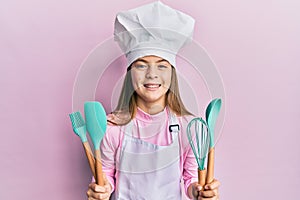 Beautiful brunette little girl wearing professional cook apron holding cooking tools smiling and laughing hard out loud because