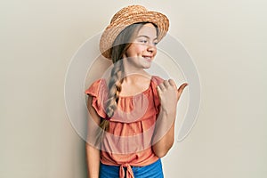 Beautiful brunette little girl wearing summer hat smiling with happy face looking and pointing to the side with thumb up