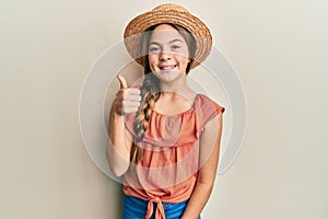 Beautiful brunette little girl wearing summer hat smiling happy and positive, thumb up doing excellent and approval sign