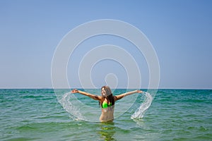 Beautiful brunette with long hair stands waist-deep in the ocean and splashes her hands in water. Young slender girl in ocean